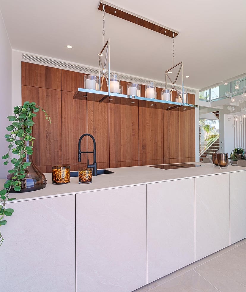 Modern kitchen island with matte white surfaces and dark wood wall-to-ceiling cabinetry, interior glass staircase visible in background.