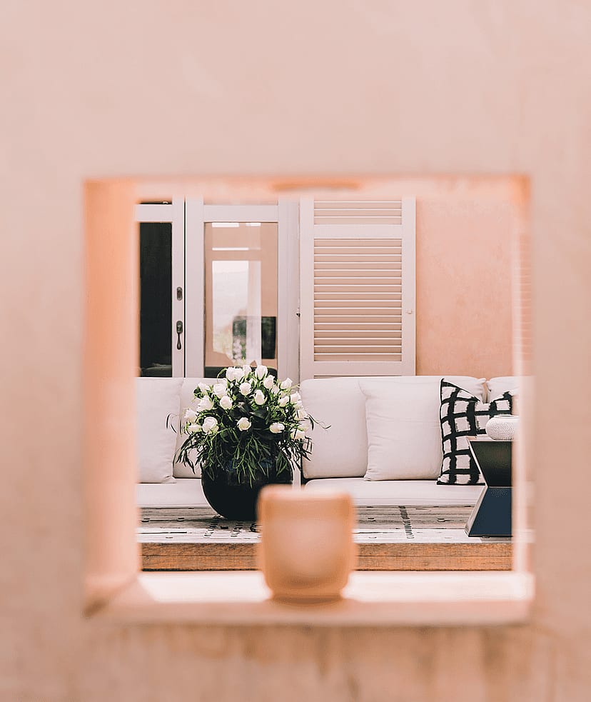 A square architectural opening in a pink stucco wall reveals a white sofa with patterned pillows and a potted plant on a terrace.