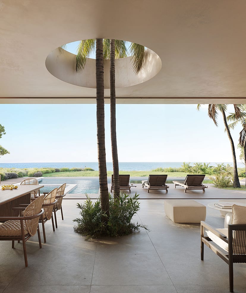 A wide outdoor covered terrace featuring a dining area, lounge seating, and an open circular skylight with trees, facing the sea.