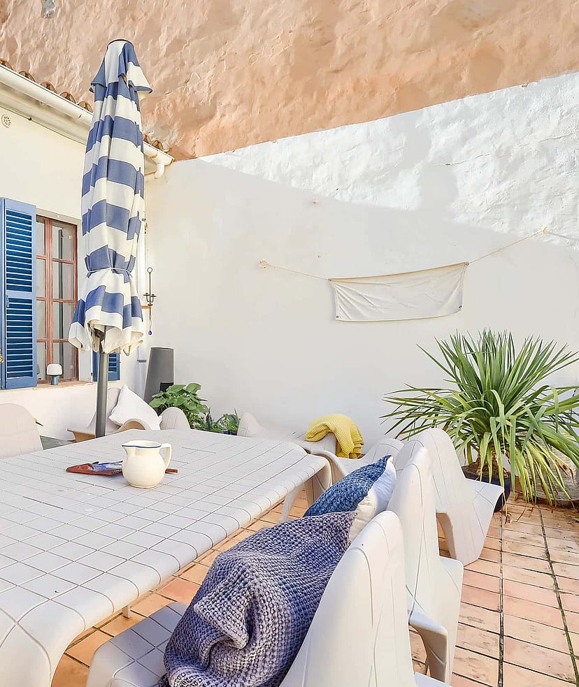Outdoor terrace with a dining table and chairs, blue-striped sun umbrella, and blue shutters on a white building exterior.