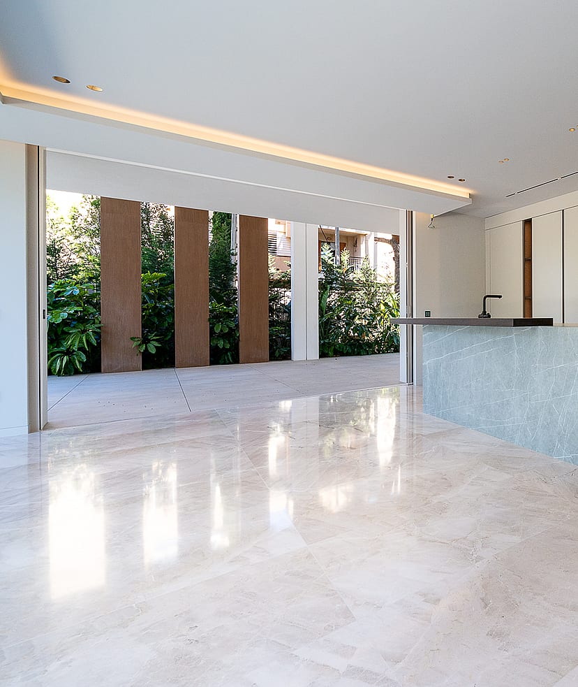 Modern kitchen featuring a large grey stone island, handleless white cabinets, and polished light marble floors. A wide glass door opens to a stone terrace and garden.