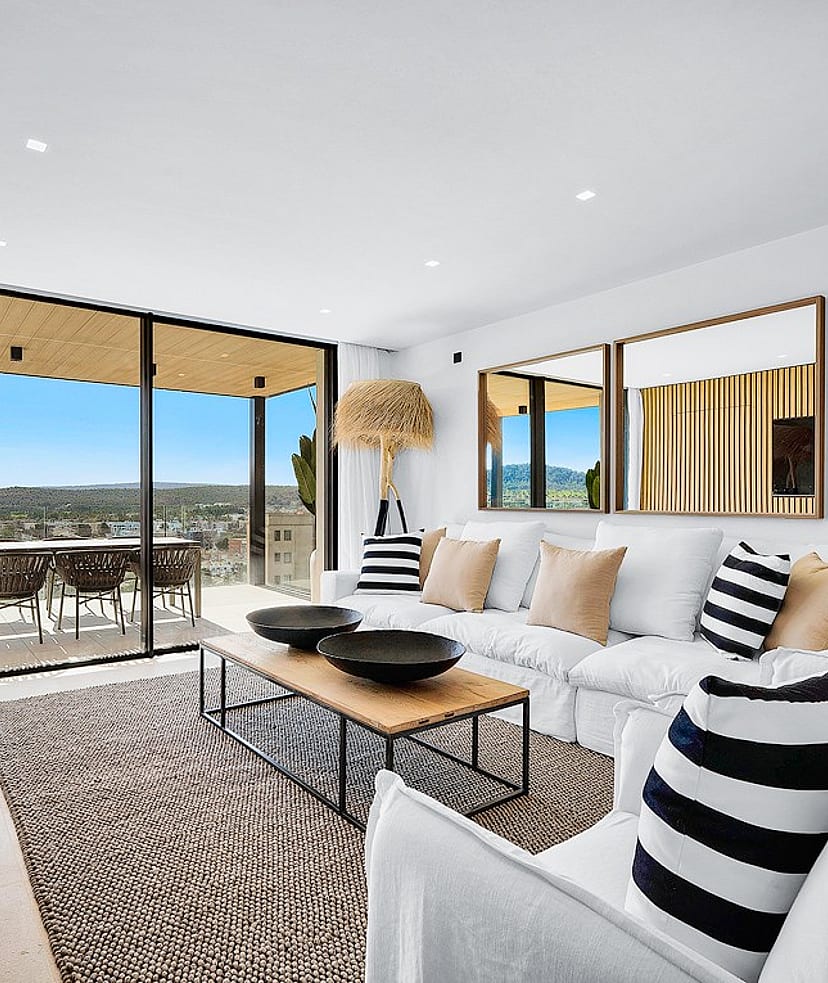 Modern open-plan living room with white sofas, wooden wall paneling, and large glass doors opening onto a terrace with mountain views.
