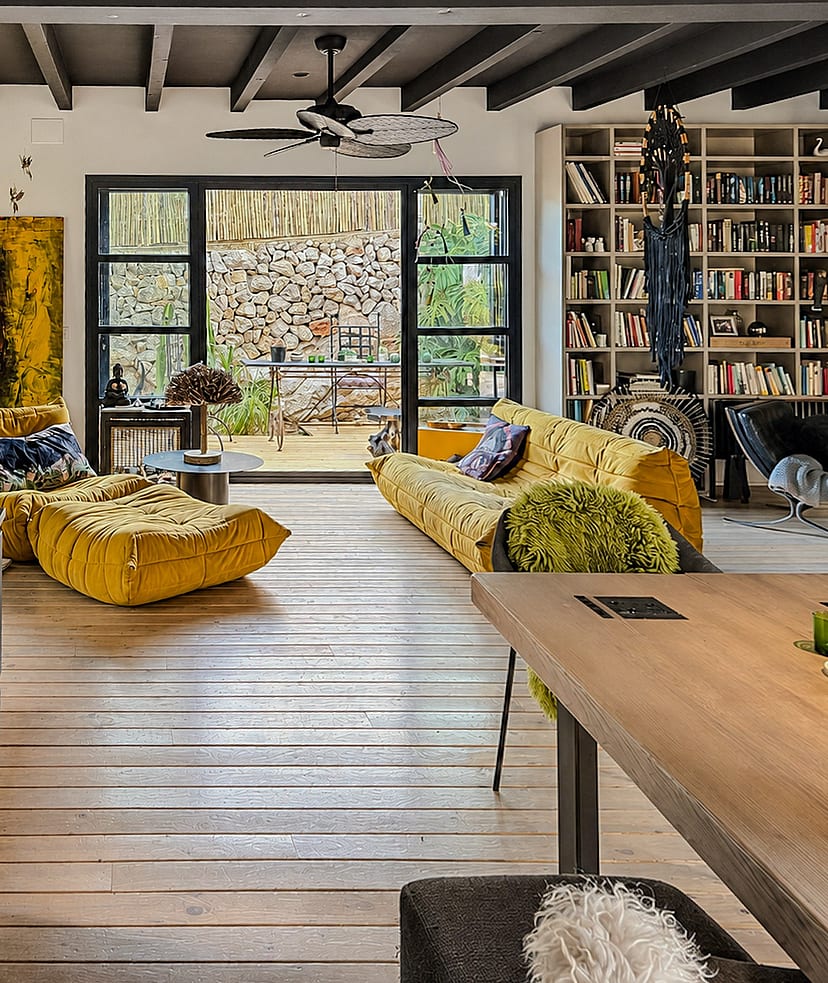A living area with light wood floors, a large yellow sofa, and a wall-to-wall integrated bookshelf opening onto a stone terrace.