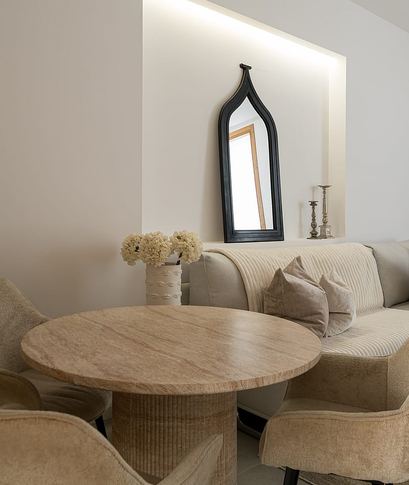 A round travertine stone dining table set in a living room with a beige textured sofa and a black arched mirror on the wall.