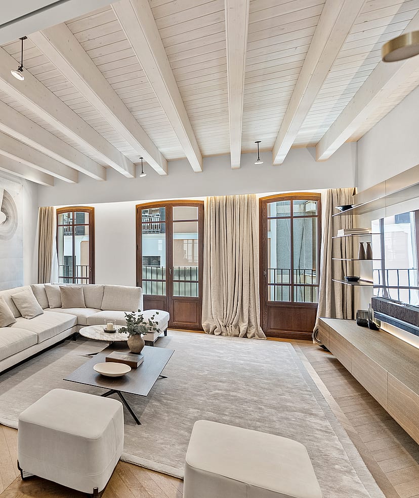 Bright living room with herringbone wood flooring, white sectional sofa, and tall wooden French doors under a beamed ceiling.