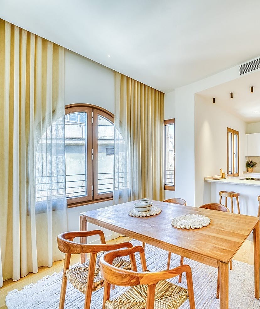 A bright open-plan kitchen and dining room with wooden furniture, white cabinetry, and large arched windows draped in curtains.