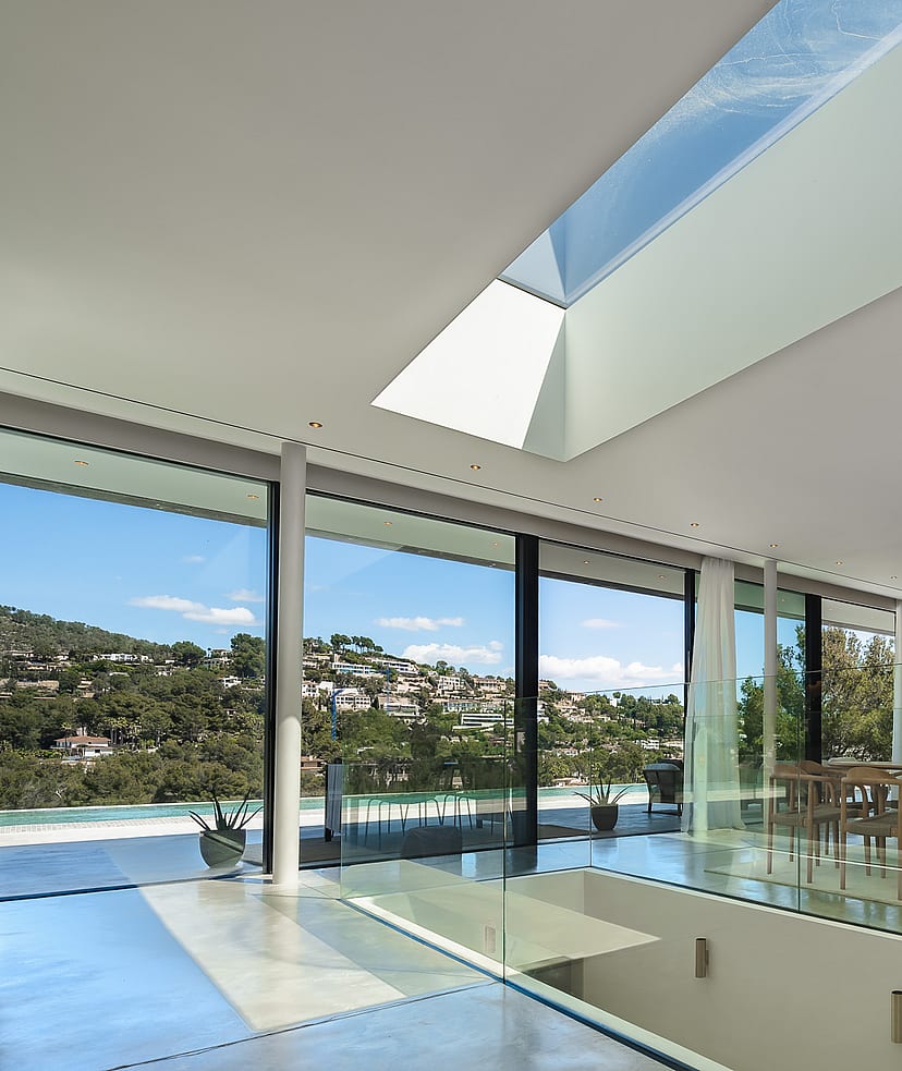 Modern living and dining room featuring floor-to-ceiling glass walls, a large skylight, and views of Son Vida hills.