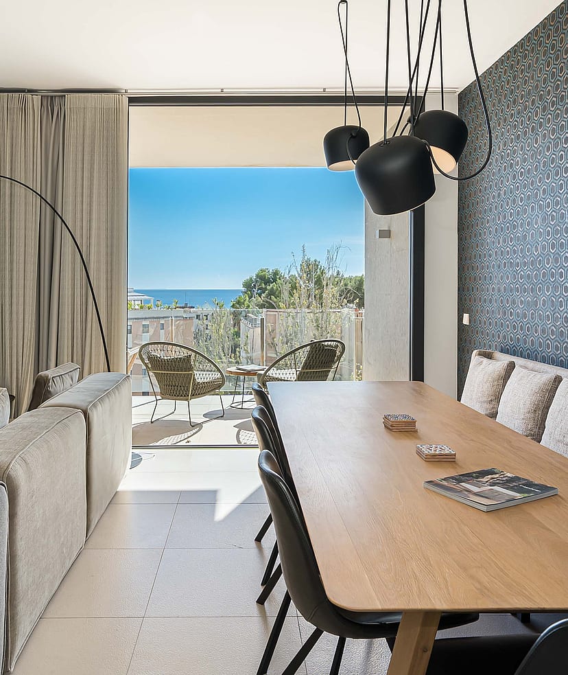 Interior view of an open-plan living and dining room with floor-to-ceiling glass doors opening onto a terrace with a sea view.