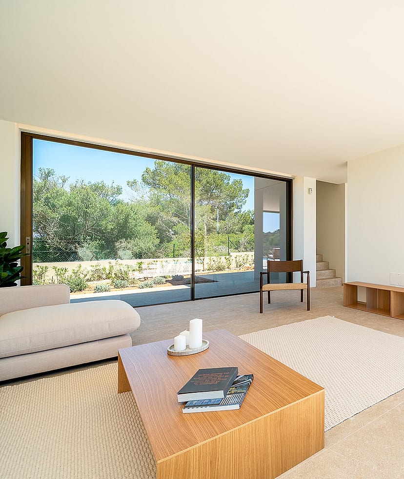 A bright, open-plan living room with a light beige sofa, wooden coffee table, and large sliding glass doors overlooking trees.