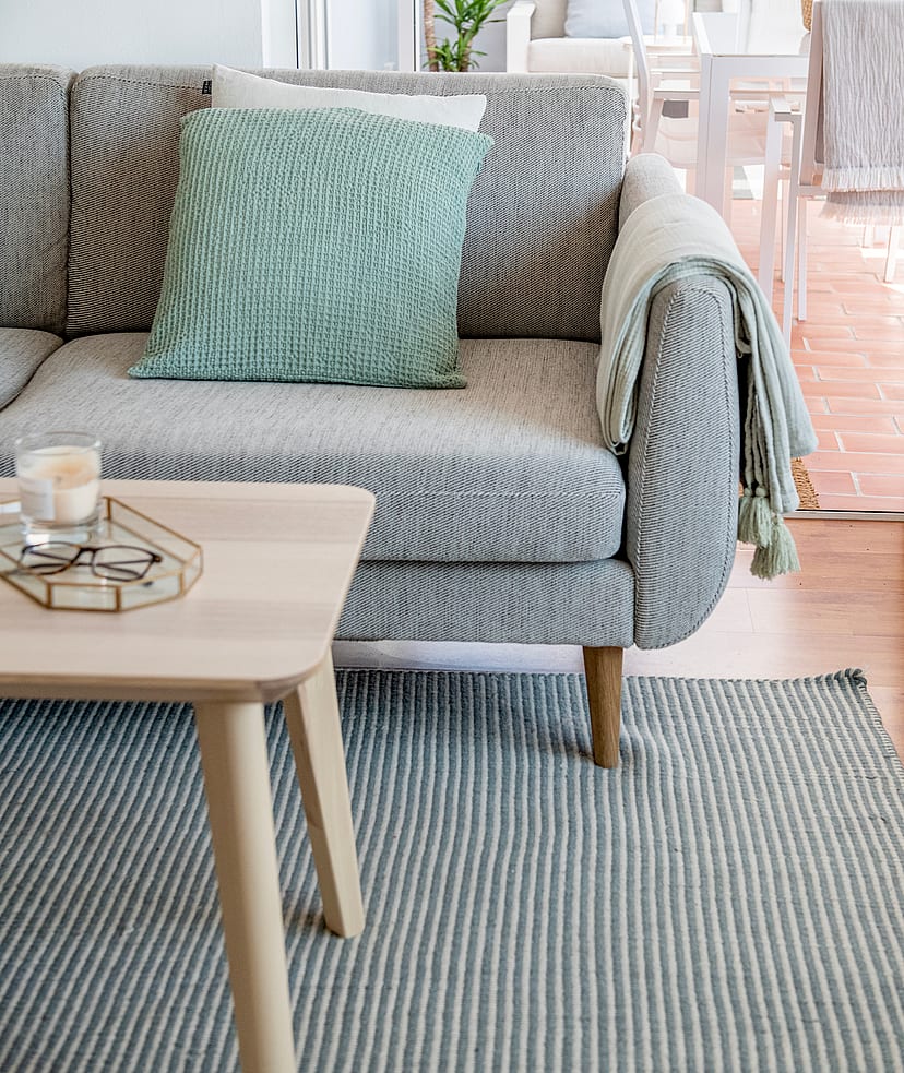 Close-up of a grey woven sofa with a mint green cushion and a light wood coffee table on a striped rug, leading to a terrace.