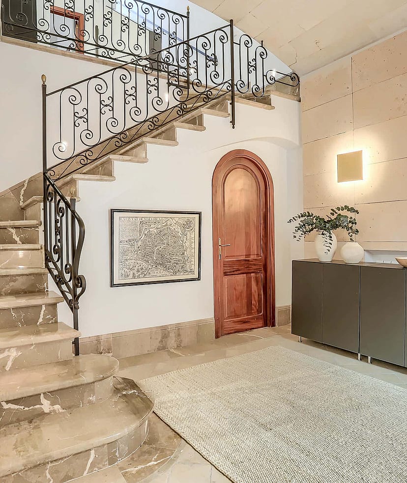 An interior shot of an entrance hall featuring a wide marble staircase with a wrought-iron railing, a curved wooden door, and a textured stone feature wall.