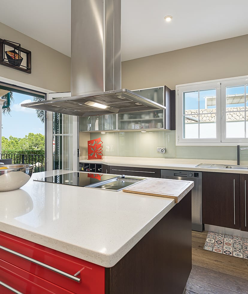 A modern kitchen featuring a bold red central island with a white stone countertop, dark cabinetry, and floor-to-ceiling glass doors opening to a terrace.