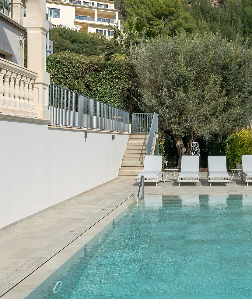 An outdoor rectangular swimming pool with four white loungers on a stone-paved terrace, bordered by a white villa wall.