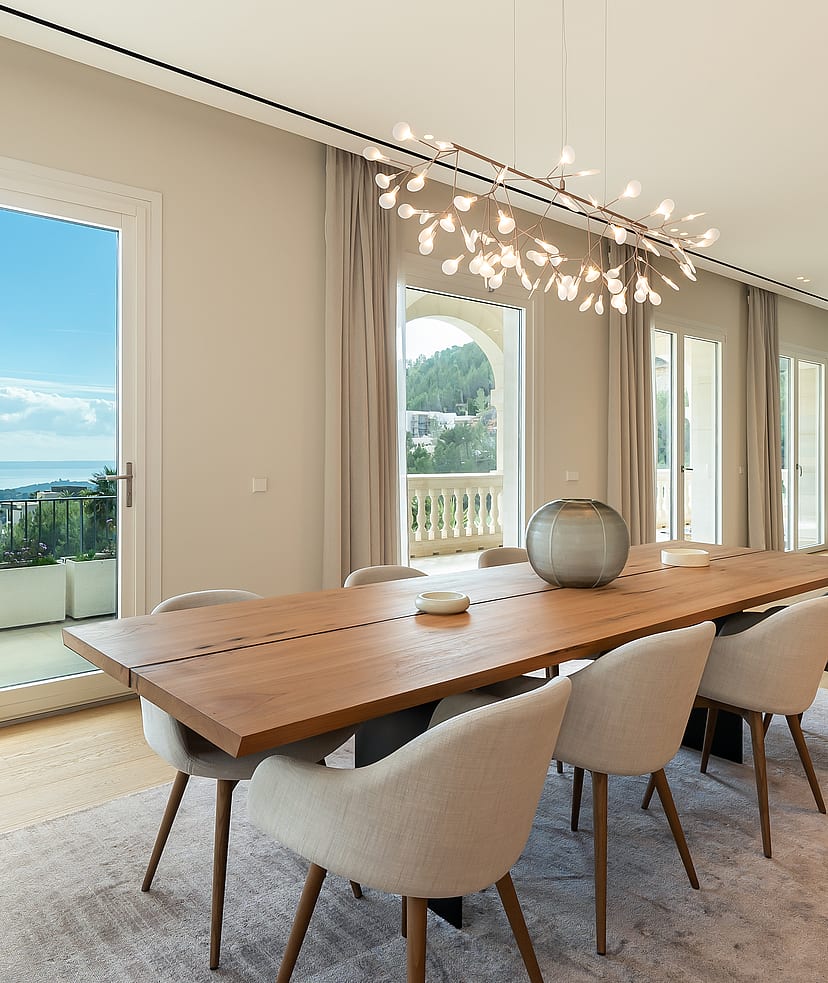 Dining room featuring a long wooden table, light-colored upholstered chairs, and a sculptural chandelier, overlooking the sea.