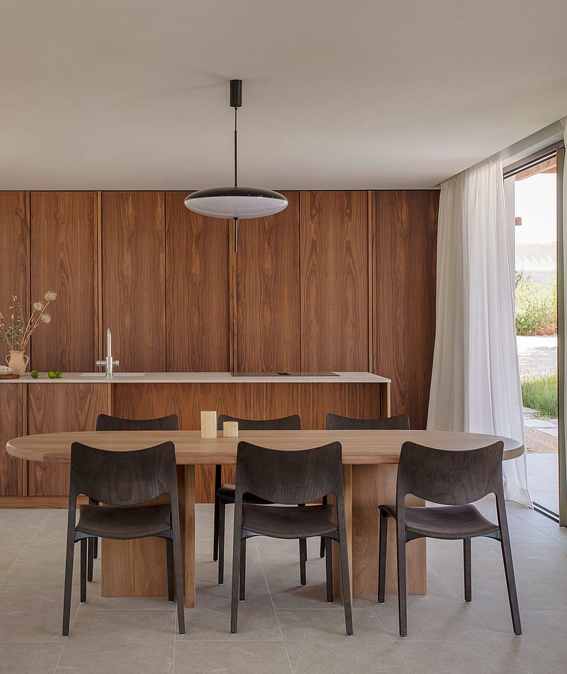 A minimalist dining area featuring an oval wood table and dark chairs, set against full-height walnut kitchen cabinetry.