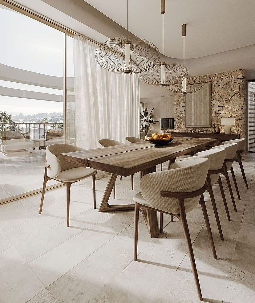 Dining area with long wooden table, cream upholstered chairs, and a natural stone feature wall, opening onto a balcony.