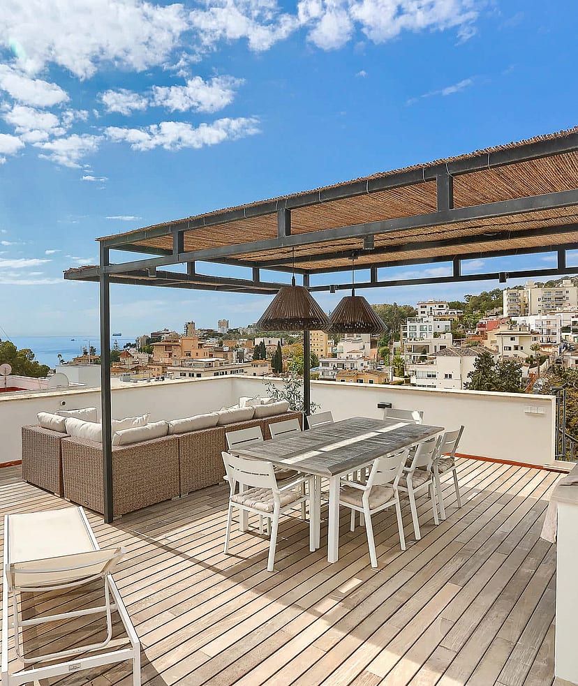 A spacious outdoor rooftop terrace featuring a wooden deck, a covered dining area with a woven pergola, lounge seating, and distant sea views over a coastal neighborhood.