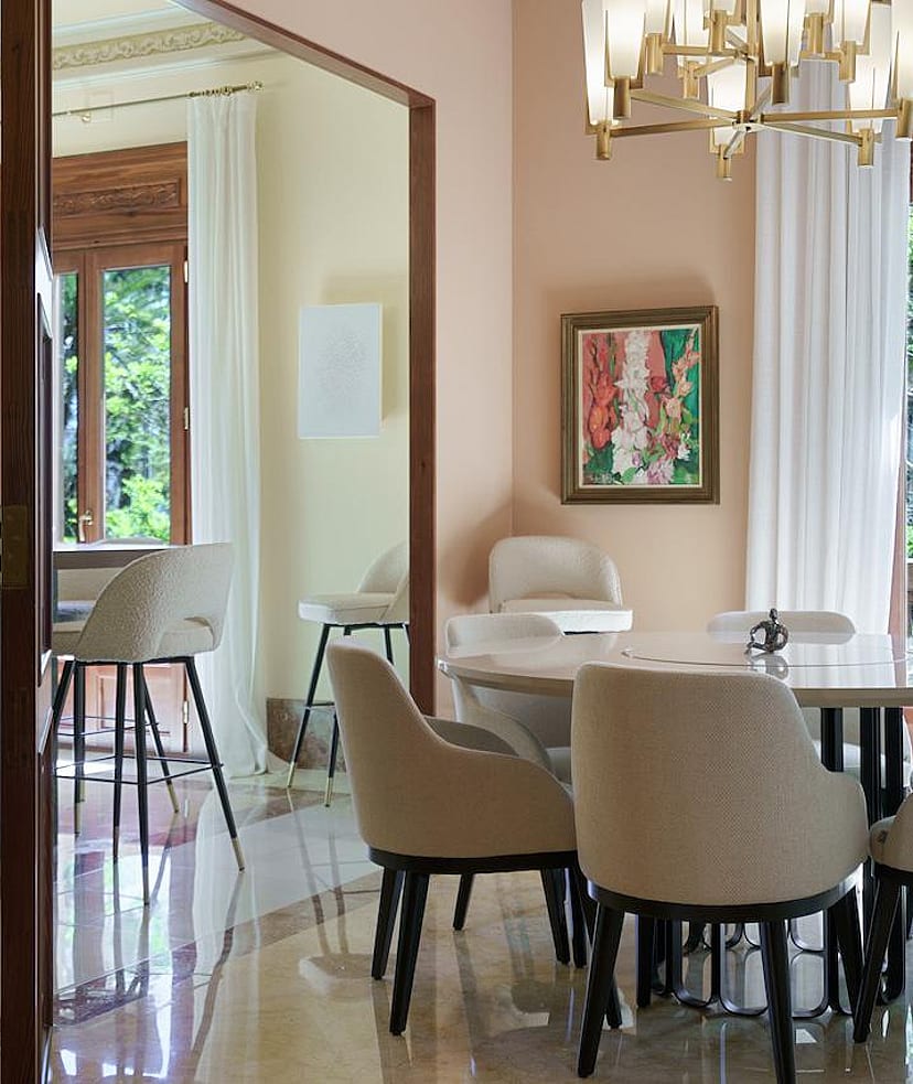 Dining room with a round marble table, upholstered chairs, a crystal chandelier, and large windows overlooking trees.