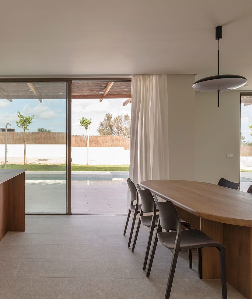 An oval oak dining table and dark chairs in a sunlit room with stone floors and floor-to-ceiling glass sliding doors.