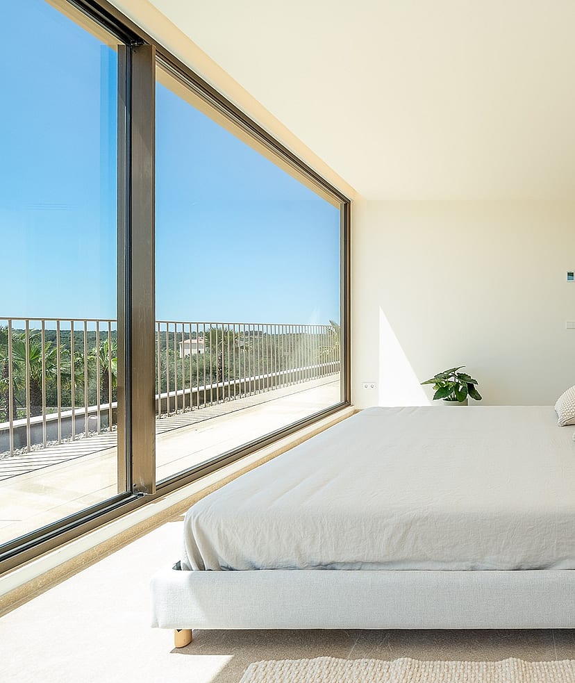 Modern bedroom with a double bed, floor-to-ceiling glass wall, white walls, and view of the exterior landscape.