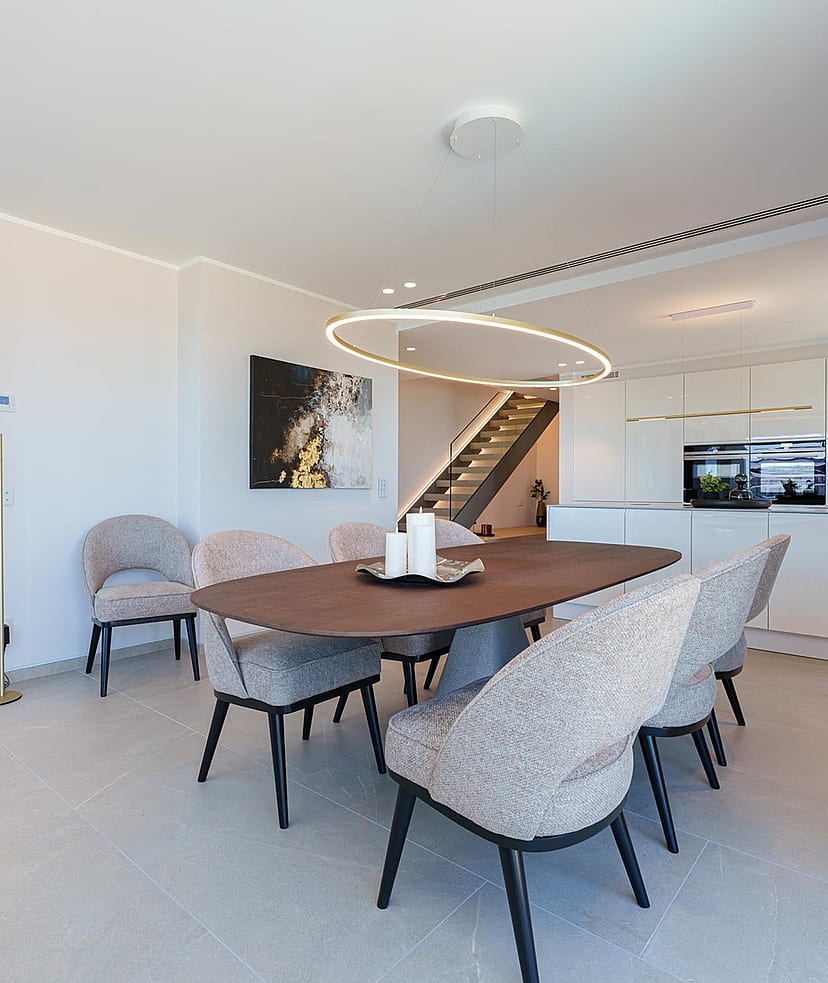 Comedor con mesa ovalada de madera y seis sillas, junto a una cocina blanca minimalista con iluminación empotrada.