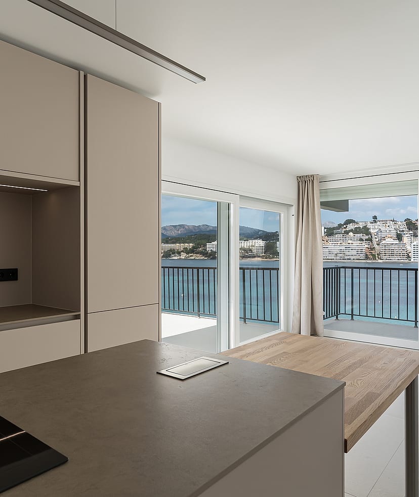 Modern taupe kitchen and wooden island extension facing floor-to-ceiling windows with a view of a coastal harbor.