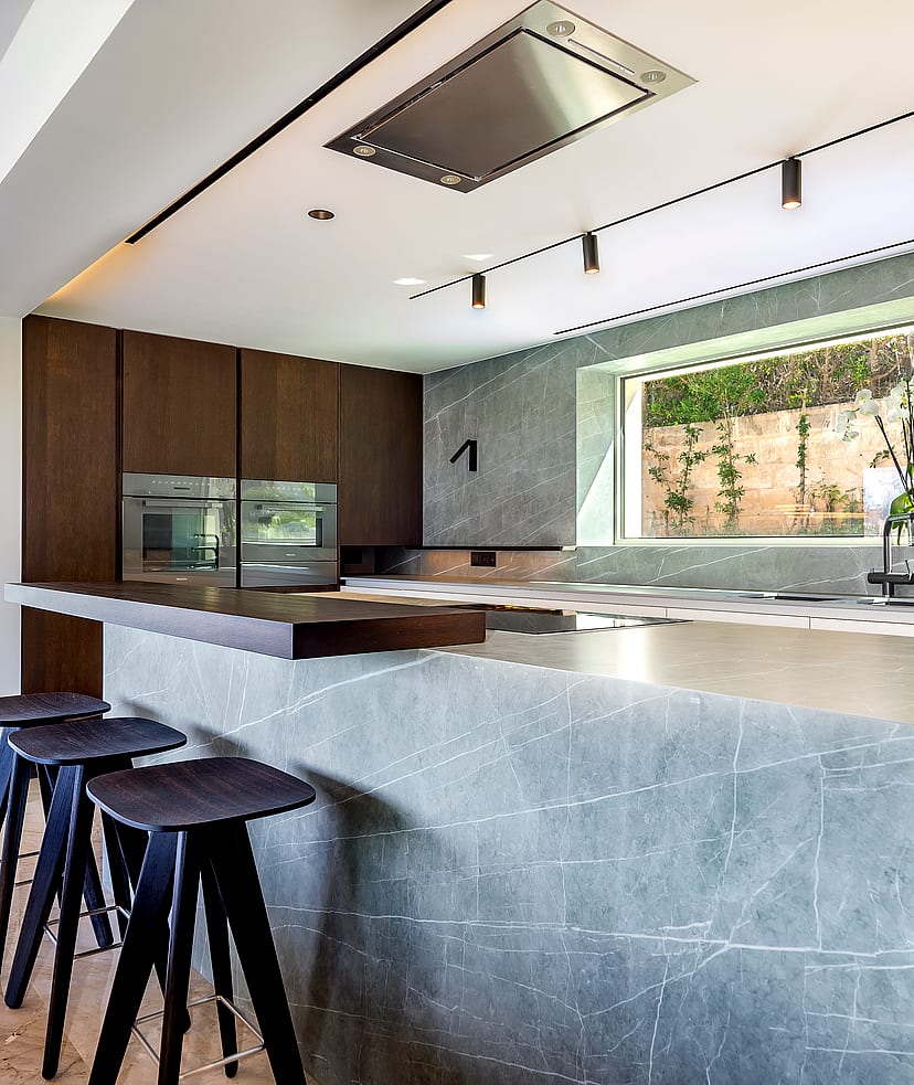 Modern kitchen with a large grey marble island, wooden cabinets, and three dark wooden bar stools under track lighting.