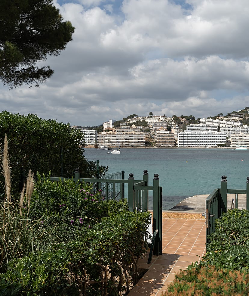 A view through a dark green garden gate over a terracotta terrace toward a calm turquoise bay and coastal buildings.