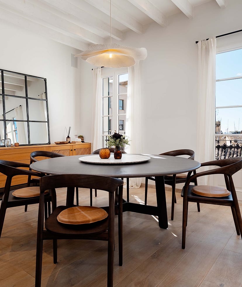Round dark wood dining table with woven-seat chairs beneath a white beamed ceiling, French doors opening to rooftop views.