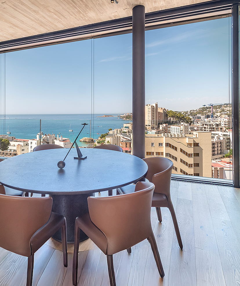 Round dark wood dining table surrounded by leather chairs next to floor-to-ceiling windows overlooking the sea and coastline.