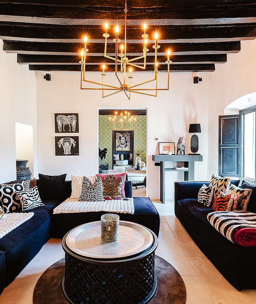 Interior view of a living room with dark low-profile sofas, a central round metal coffee table, and an ornate brass chandelier hanging from dark exposed ceiling beams.
