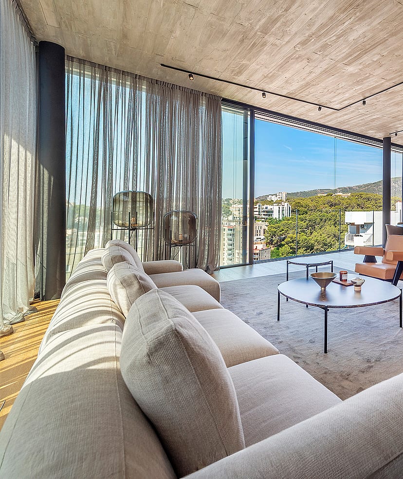 Wide-angle shot of a bright, open-plan living room with exposed concrete ceilings, light wood floors, and floor-to-ceiling glass walls looking out to coastal scenery.