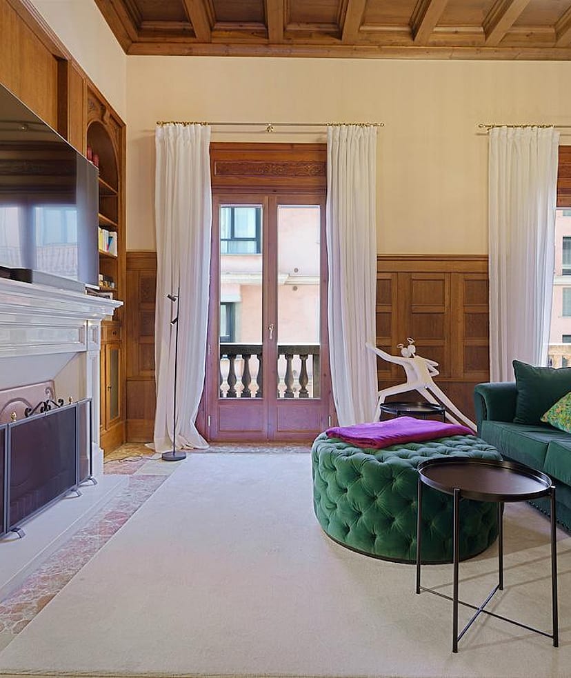 A formal living area featuring dark wood wall paneling, a stone fireplace, a green velvet sofa, and tall windows facing the city.