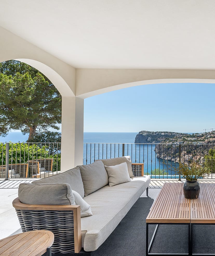 A covered outdoor lounge featuring gray sofas and wooden tables under white arches, overlooking the blue sea and cliffs.