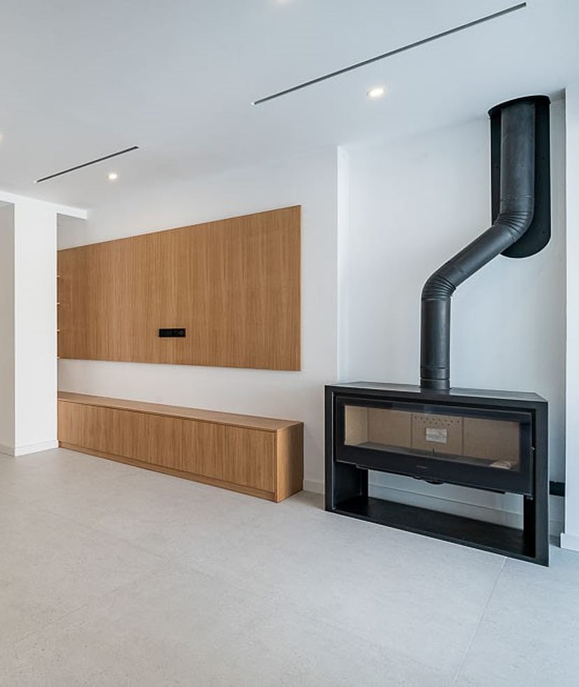 An expansive, bright living space with large light-grey floor tiles, custom light oak cabinetry, and a black metal fireplace.