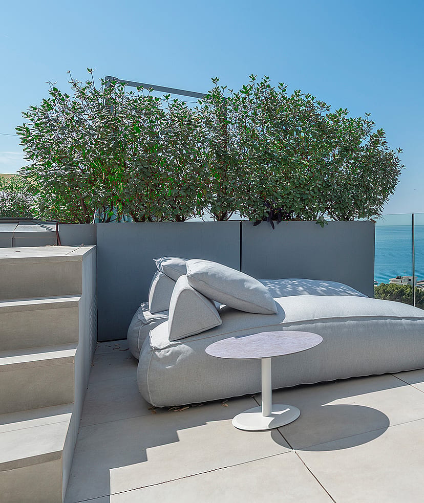 A rooftop terrace featuring a large gray outdoor daybed, a white side table, and a glass balustrade overlooking the sea.