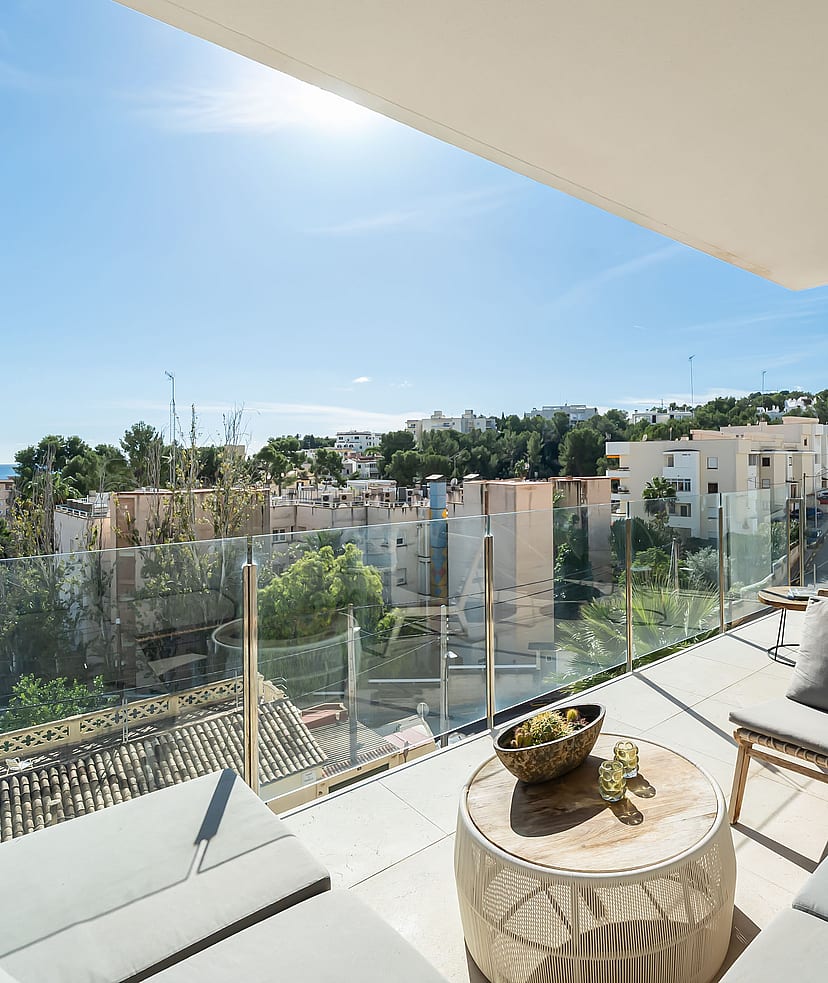 A covered terrace with grey outdoor sofas, a circular wooden coffee table, and glass railings overlooking a coastal town.