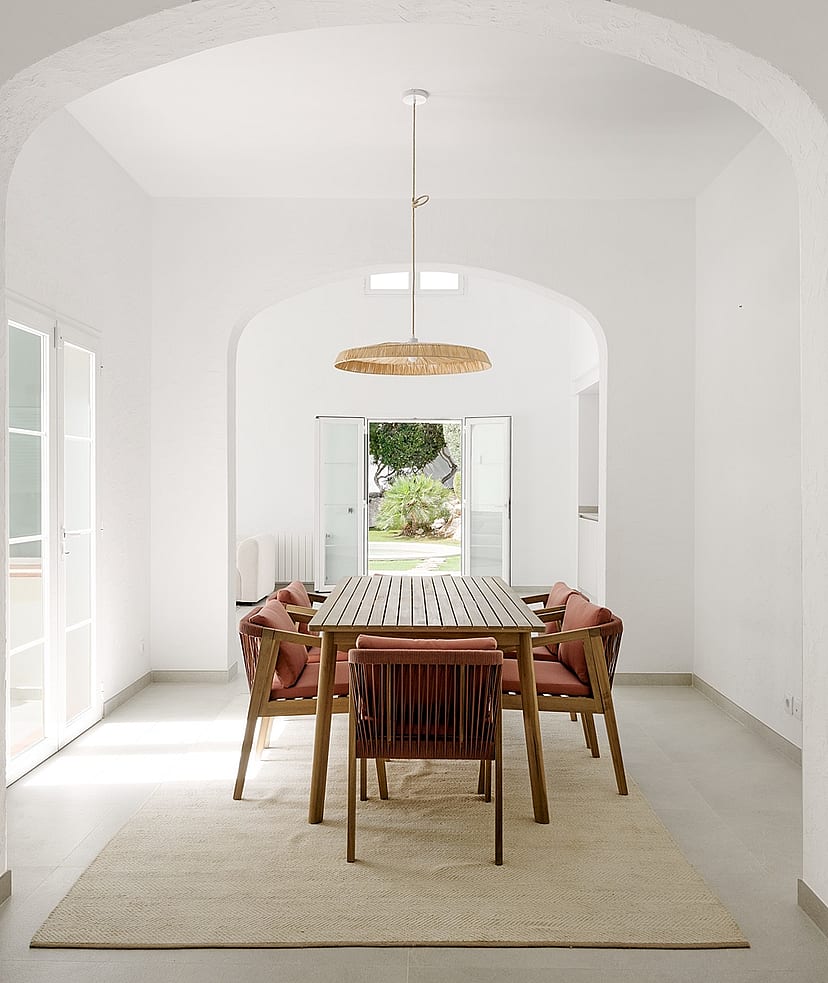 Comedor con bóveda blanca, mesa de teca, sillas de cuerda y lámpara de ratán, vistas al jardín a través de puertas abiertas