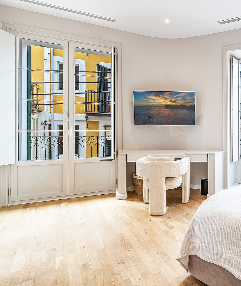 Bright bedroom with white walls, light wood floors, a desk, and floor-to-ceiling windows looking out onto a yellow building.