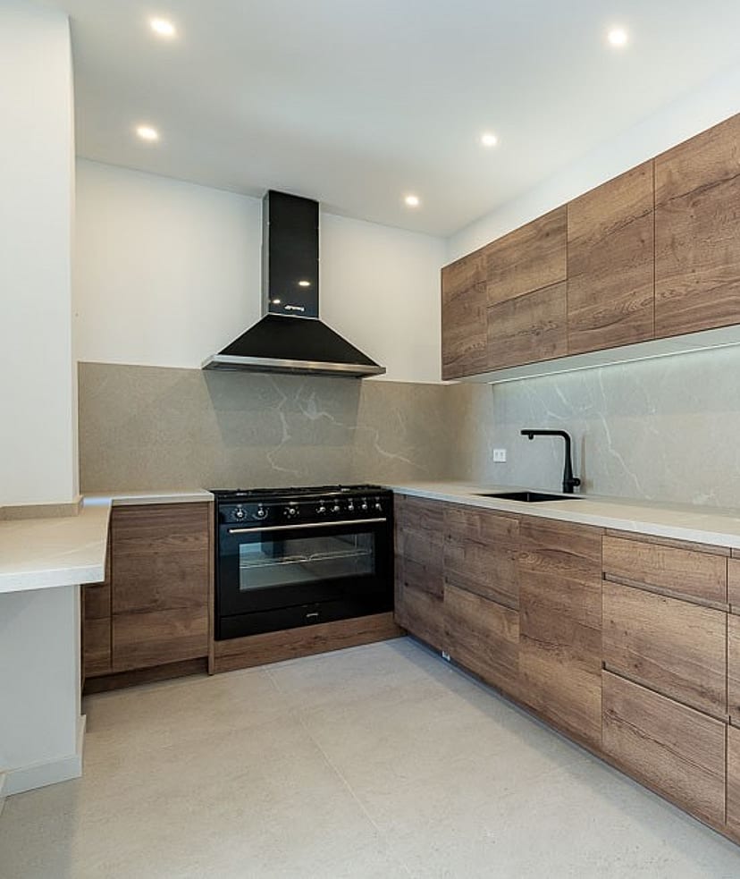A modern kitchen in a townhouse featuring wood grain cabinets, dark appliances, stone countertops, and recessed ceiling lights.