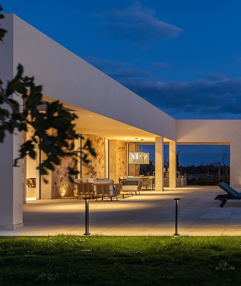 Exterior view of a modern finca at twilight, featuring a lit swimming pool, stone terrace, and warm architectural lighting.