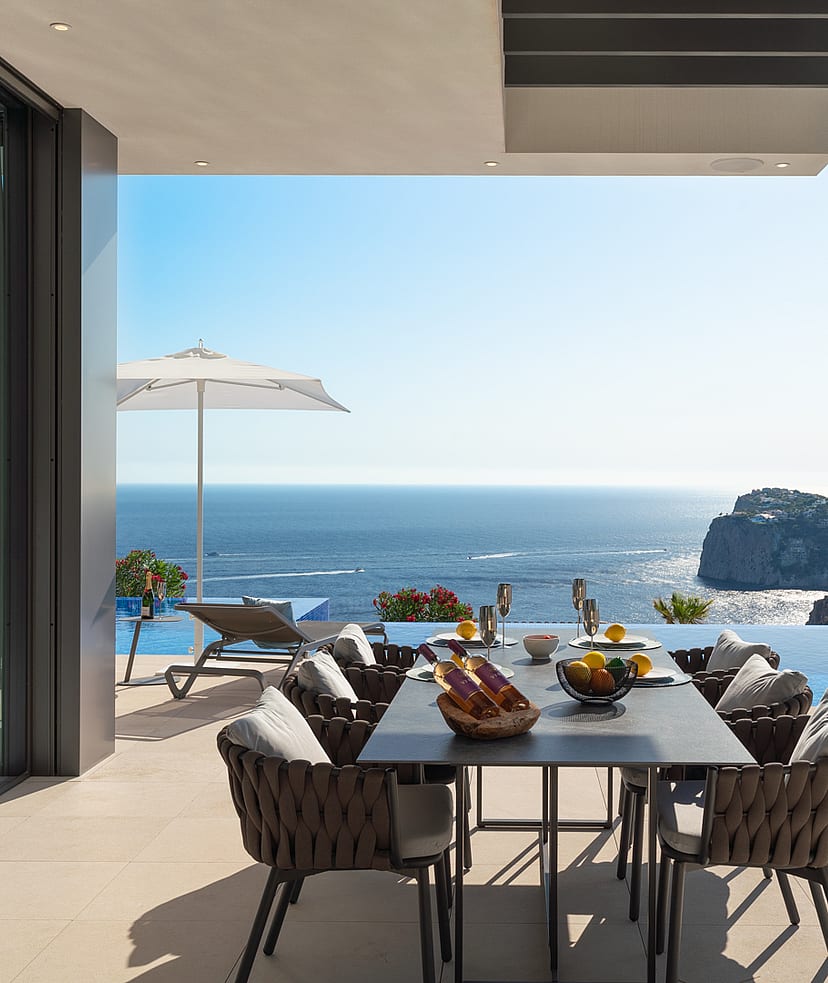 Covered outdoor dining terrace with a dark metal table, woven chairs, and an infinity pool edge against a Mediterranean sea view.