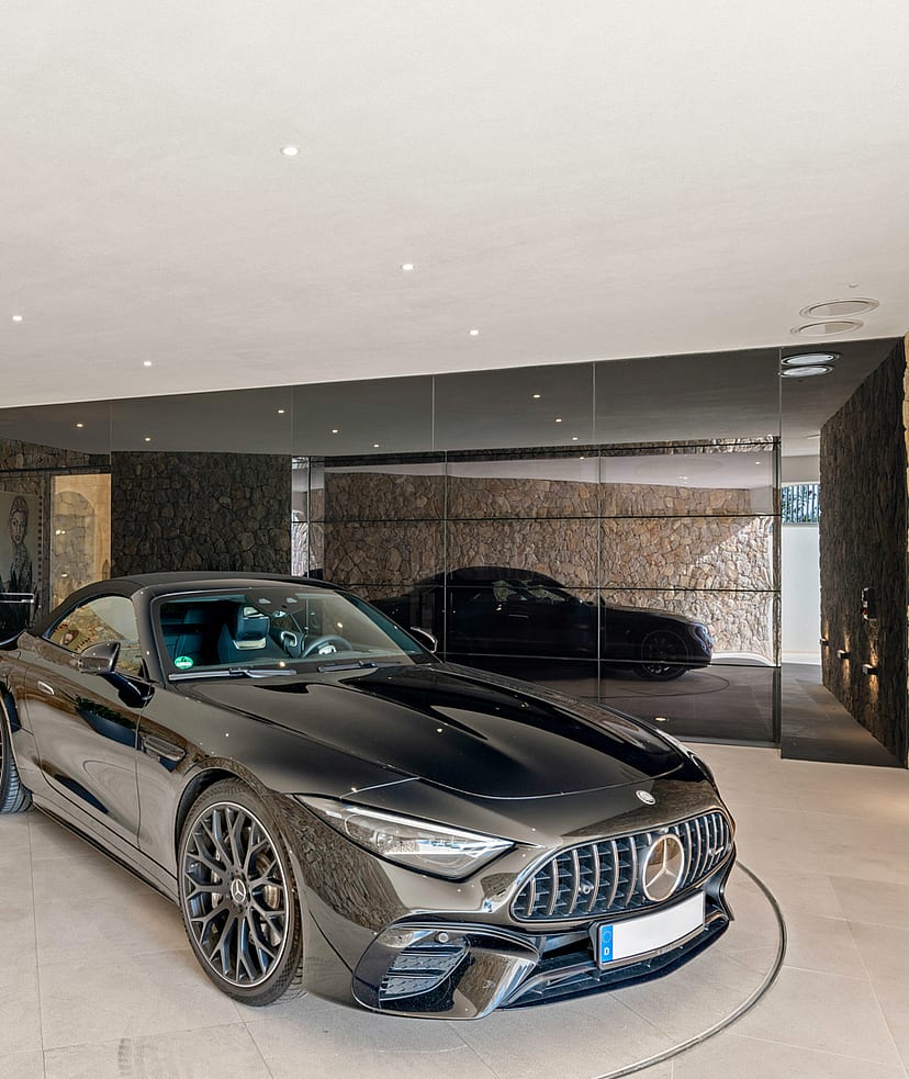 A luxury garage interior featuring a dark sports car on a mechanical turntable, flanked by natural stone walls and planters.