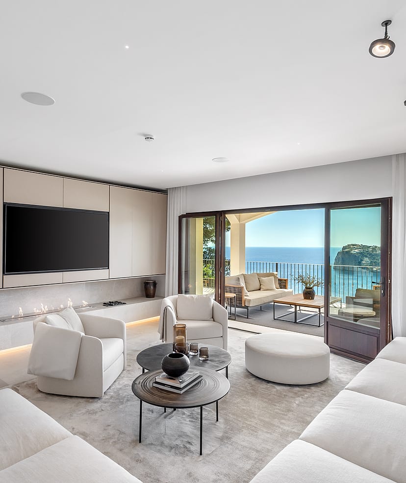 Modern white living room with soft seating and circular tables facing wide glass doors and a coastal view.