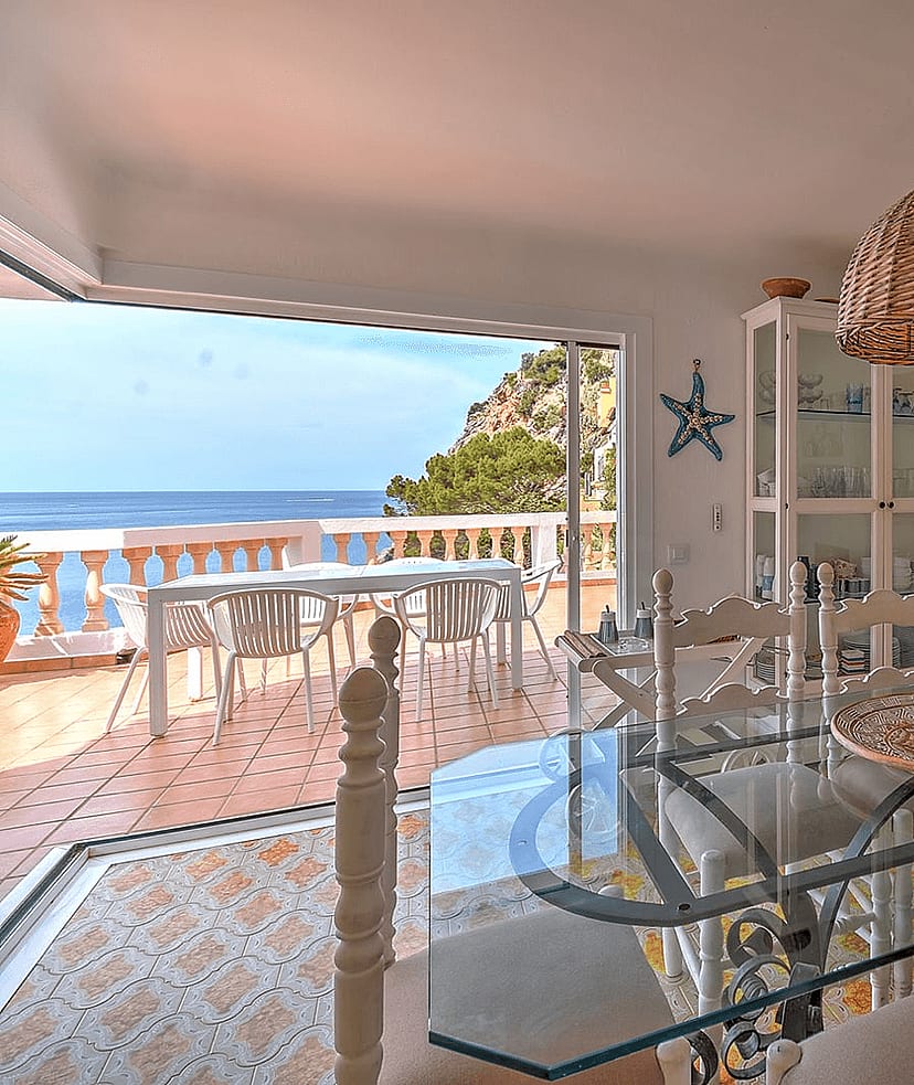 Interior dining room with a glass-top table, white wooden chairs, and a rattan pendant light, opening to a terrace with sea views.