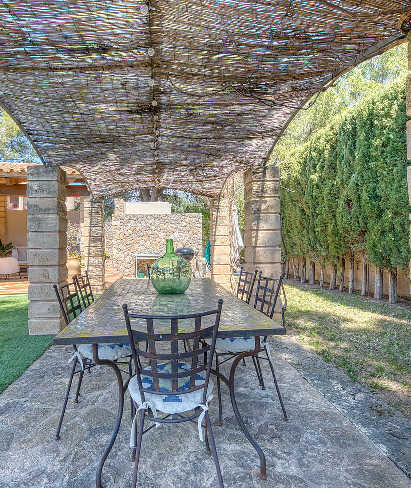 A stone-columned patio with a trellis roof and iron dining set, overlooking a green garden and building exterior.