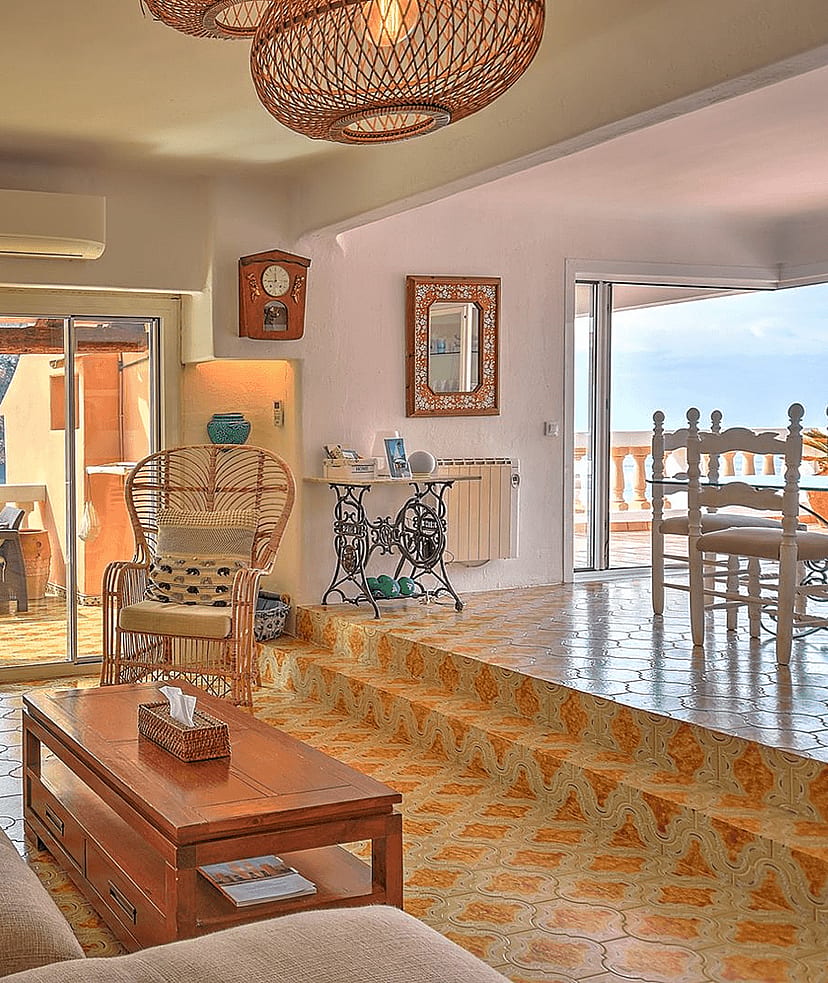 Sunlit living area with tiled floors, rattan armchairs, and a view of Cala Llamp through glass sliding doors.
