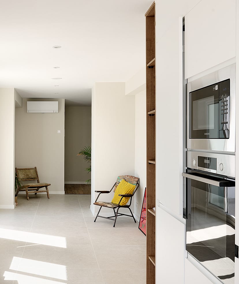 Interior view from a kitchen looking towards a living area with white walls, tiled floors, a stool, and a yellow chair.