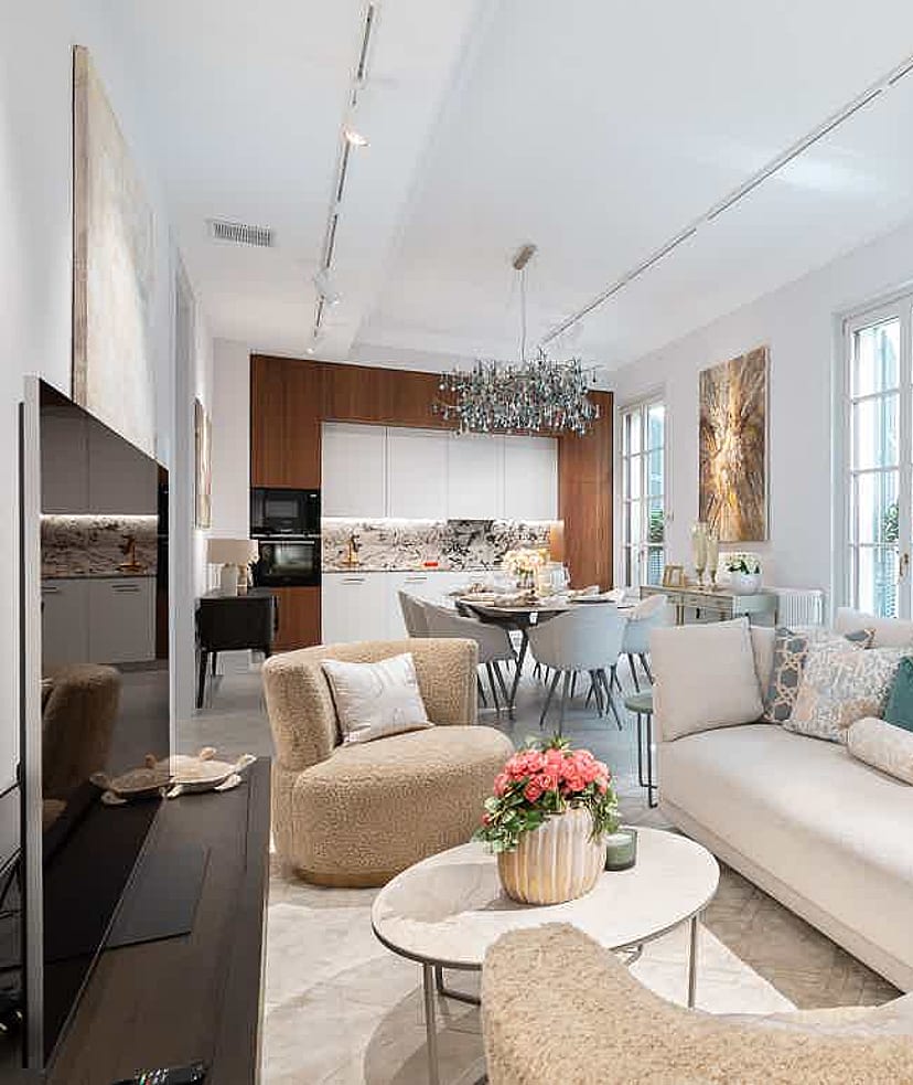 Open-plan living room with white custom built-in shelving, a neutral beige sofa, and two plush armchairs facing a kitchen area.
