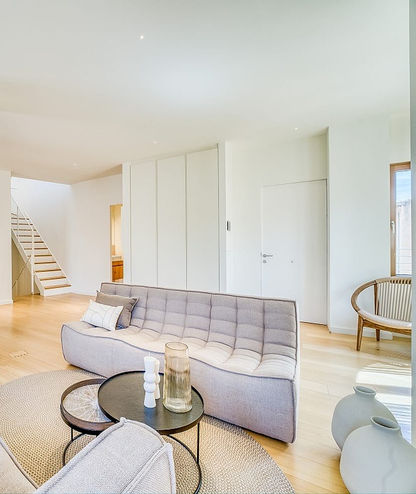 Modern open-plan living room with light oak flooring, grey tufted sofas, and a white architectural staircase.